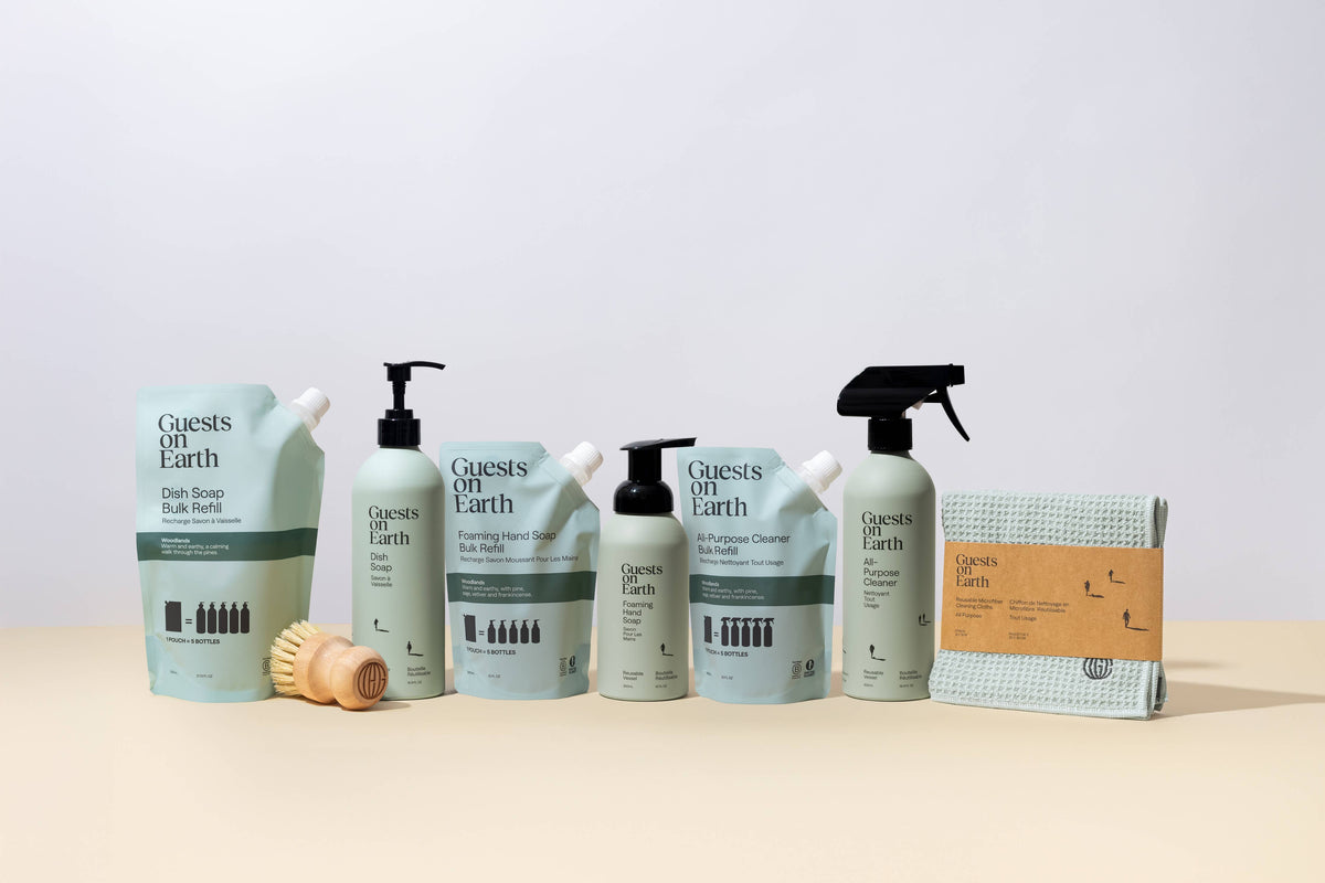 A set of Guests of Earth products arranged on a light beige surface against a white studio background. The products include a refill pouch, a pump bottle, a squeeze bottle, a spray bottle, and a packaged cloth, all in a coordinated pale green and white color scheme with black text and pump/spray details.