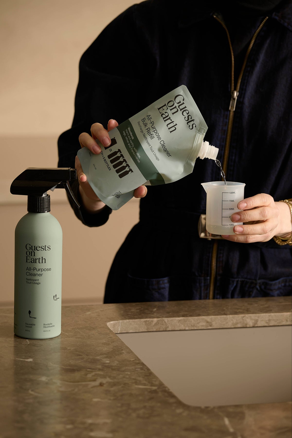 A person pours liquid from a green and white 'Guests Off Earth' all-purpose cleaner refill pouch into a clear measuring cup, with a matching green spray bottle standing on a stone countertop nearby.