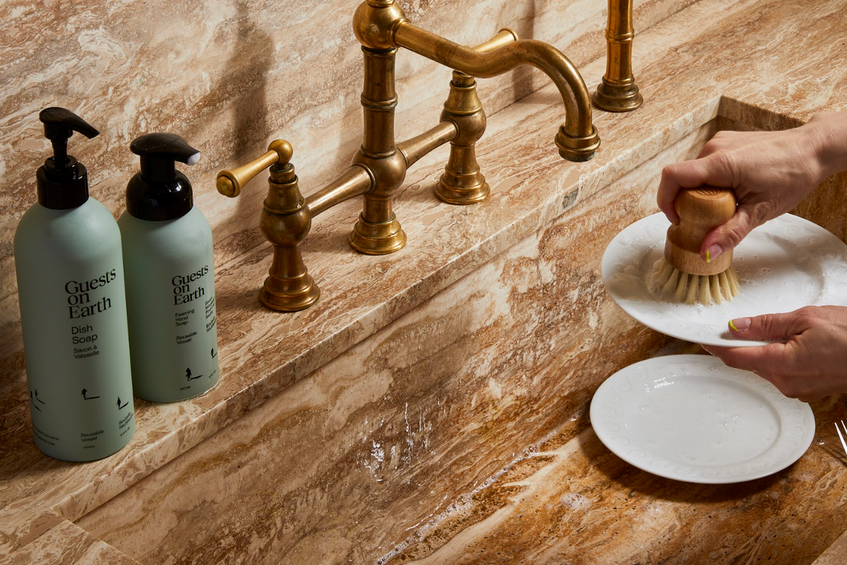 Person washing dishes with a brush using Guests on Earth dish soap, next to the bottle of soap on a marble countertop.