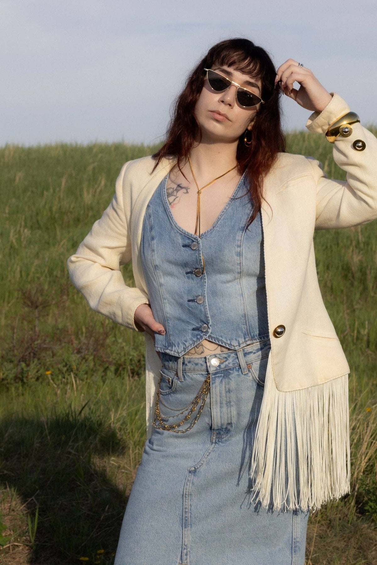 A woman wearing the Annie Sunglasses by Jenny Be Free, standing outdoors in a field. She is wearing a denim skirt and vest and a fringed jacket.
