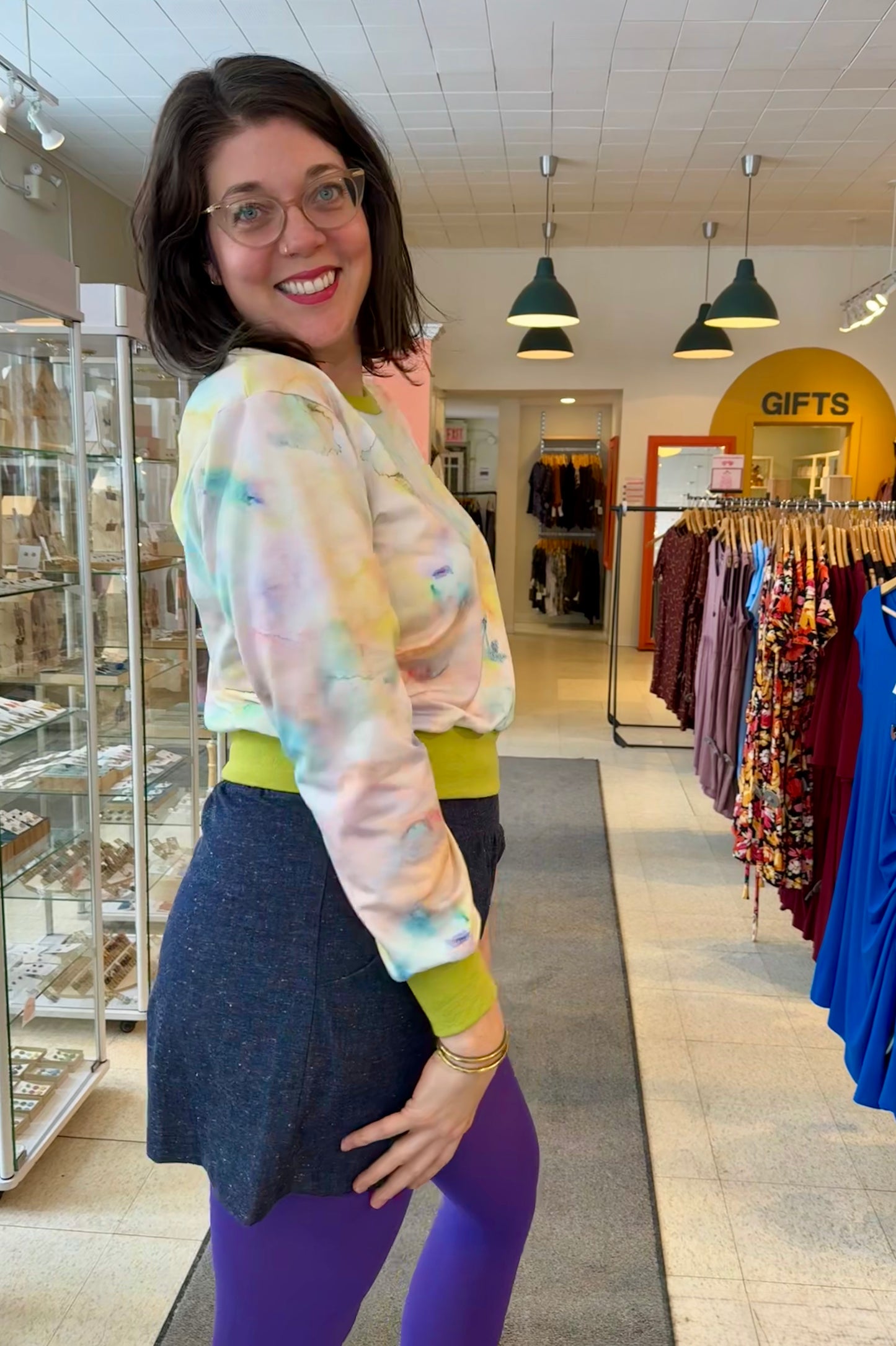 Side view of a woman wearing the Wanda E26 Sweater by Slak in Watercolour print, with a crew neck, long sleeves, and contrasting green ribbing at the cuffs, collar, and hem. She is wearing it with a navy skirt and purple tights and and standing in the store at Flock.