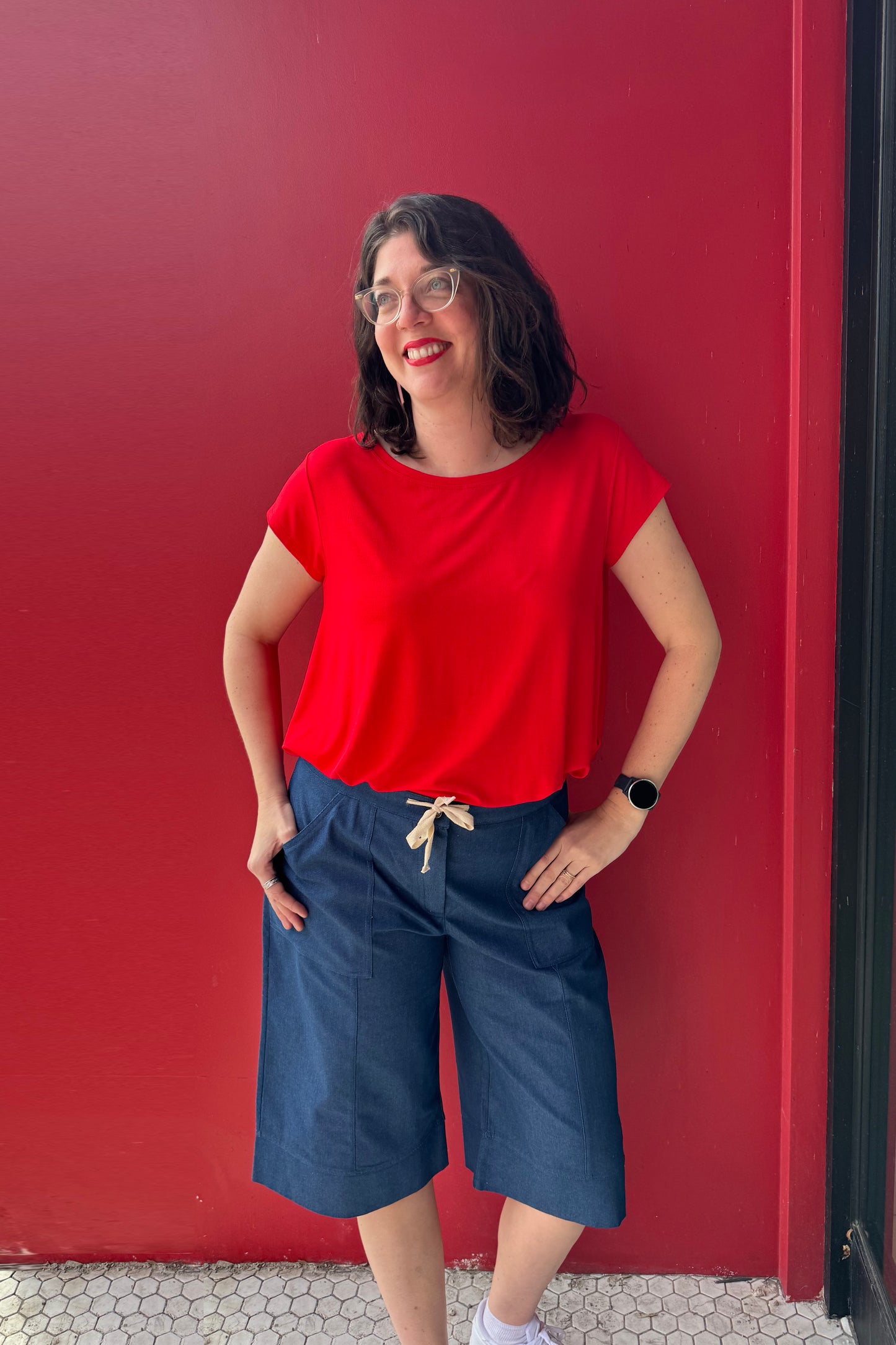 A woman wearing the Brooke Capri by Julei, wide-legged denim capris with a white drawstring and elastic at the back waist. She is wearing them with a red shirt and standing against a red wall.