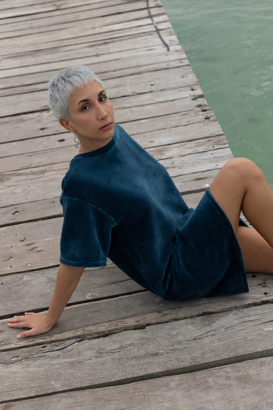 A woman wearing the Mykonos Dress by MAS in Juniper, a short sleeved boxy, knee-length dress in ribbed velvet fabric. She is sitting on a wooden dock. 