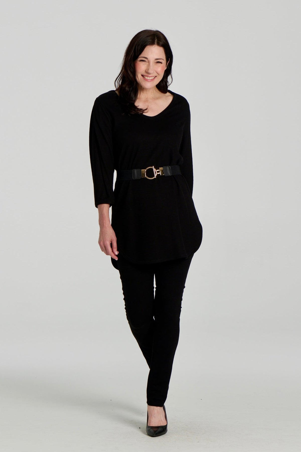  A woman wearing the Clara Tunic by Kollontai in Black, a stretch corduroy piece with a V-neck, a rounded hem, 3/4 sleeves, and inverted pleats at the shoulders. She is wearing it belted over black leggings and standing against a grey background. 