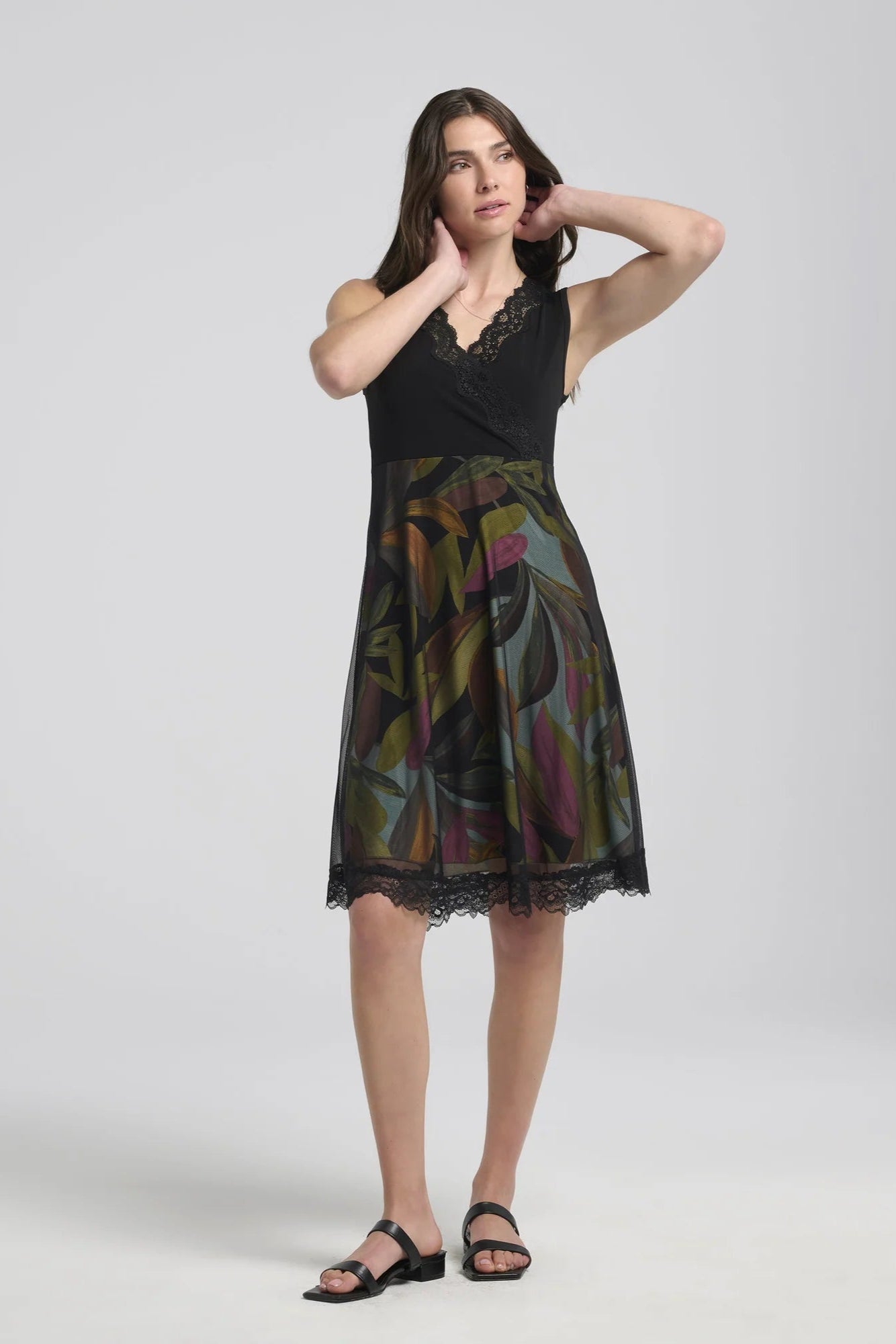A woman wearing the Deliciosa Dress by Kollontai in Black, a sleeveless dress with an empire waist, lace-trimmed cross-over neckline, and a knee-length tropical print a-line skirt with with a lace-trimmed black mesh overlay. She is standing in front of a white background. 