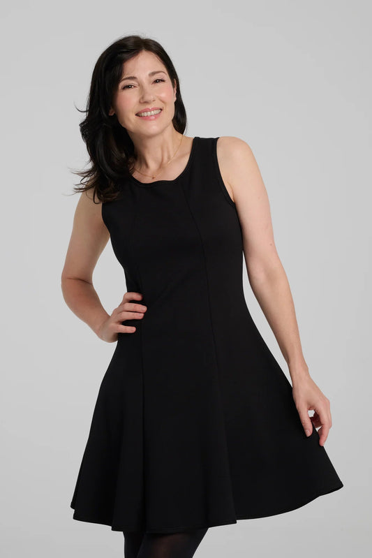 A woman wearing the Toscana Dress by Kollontai in Black, a sleeveless piece with a round neck, princess seams, and fit and flare shape with a full skirt that hits above the knee. She is standing against a grey background.
