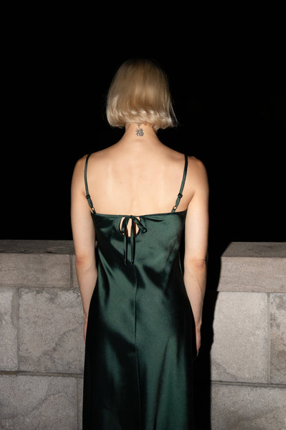 Back view of a woman wearing the Celestite Dress by Eve Lavoie in Green Satin, a slip dress with slanted spaghetti straps and an opening at the back with a tie detail. It falls to mid calf. She is standing outdoors by a stone wall. 