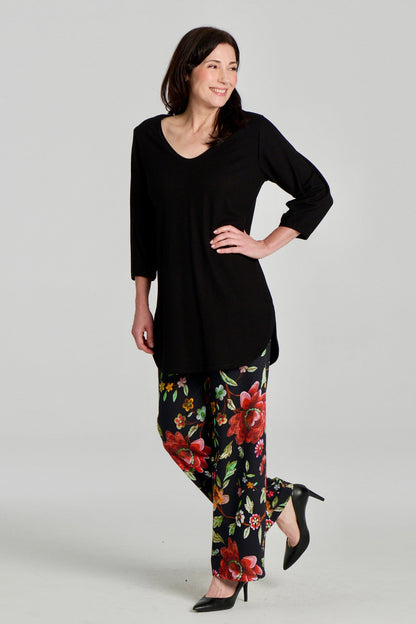 A woman wearing the Clara Tunic by Kollontai in Black, a stretch corduroy piece with a V-neck, a rounded hem, 3/4 sleeves, and inverted pleats at the shoulders. She is wearing it with the Elma Pants in floral and standing in front of a grey background.