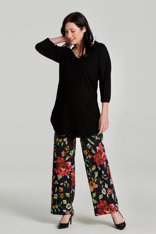 A woman wearing the Clara Tunic by Kollontai in Black, a stretch corduroy piece with a V-neck, a rounded hem, 3/4 sleeves, and inverted pleats at the shoulders. She is wearing it with the Elma Pants in floral and standing in front of a grey background.