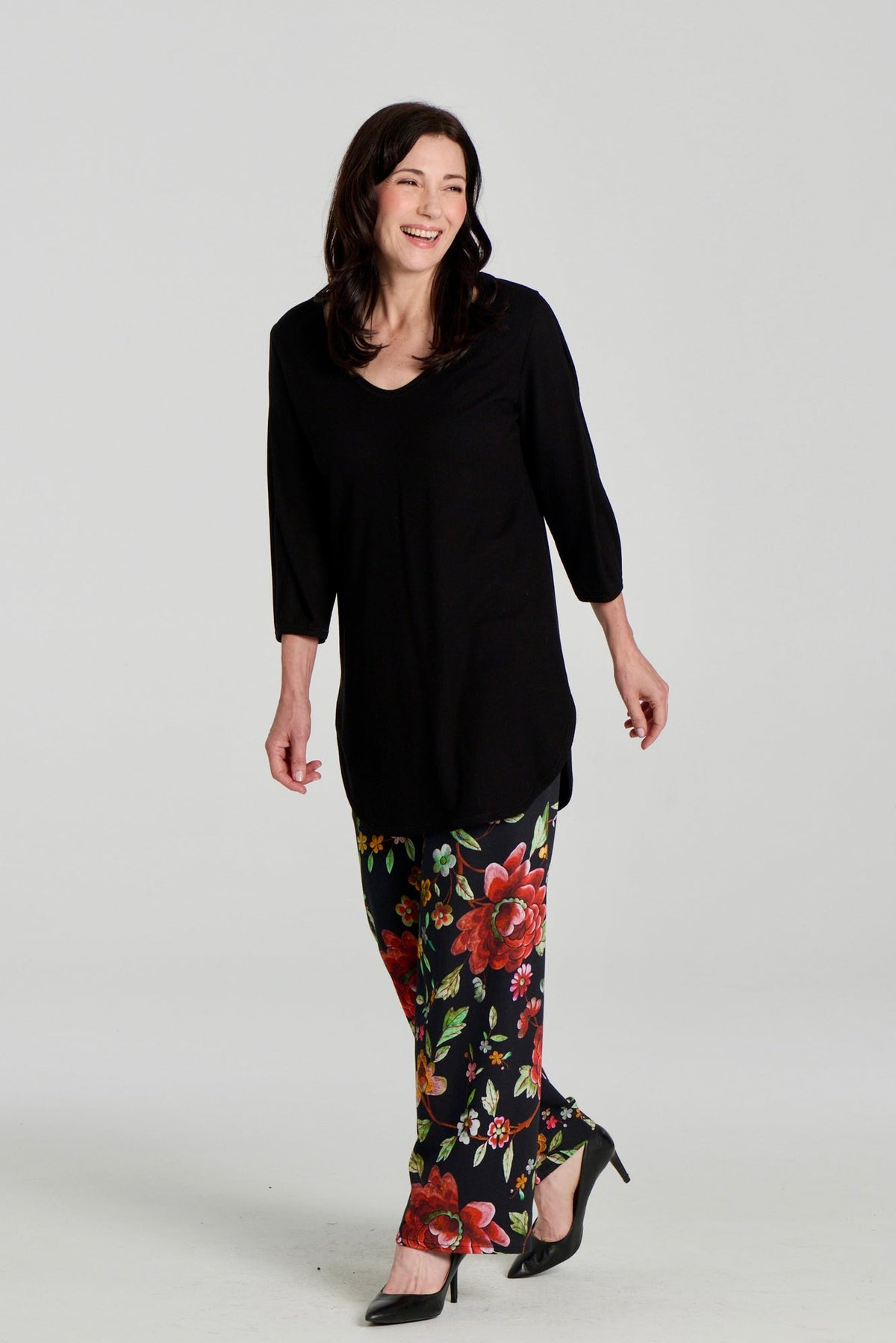 A woman wearing the Clara Tunic by Kollontai in Black, a stretch corduroy piece with a V-neck, a rounded hem, 3/4 sleeves, and inverted pleats at the shoulders. She is wearing it with the Elma Pants in floral and standing in front of a grey background.