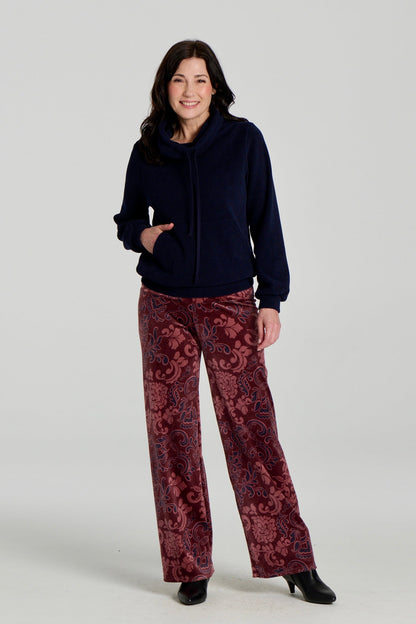 A woman wearing the Ramos Sweater by Kollontai with floral burgundy velvet pants. The Sweater is made from a navy ribbed fabric and has a large cowl neck with a drawstring and a kangaroo pocket. She is standing against a white background. 