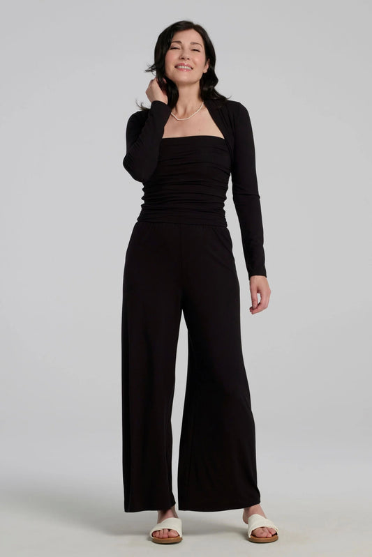 A woman wearing the Osimo Bolero by Kollontai in Black with the Dolora Jumpsuit, standing against a grey background. 