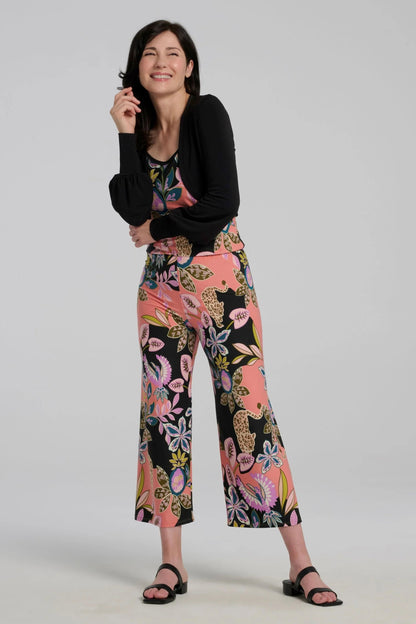 A woman wearing the Ortiga Jumpsuit by Kollontai, featuring a cheetah/floral print, spaghetti straps, an elastic waist, and 7/8 straight legs. She is wearing it with a black bolero and standing in front of a grey background.