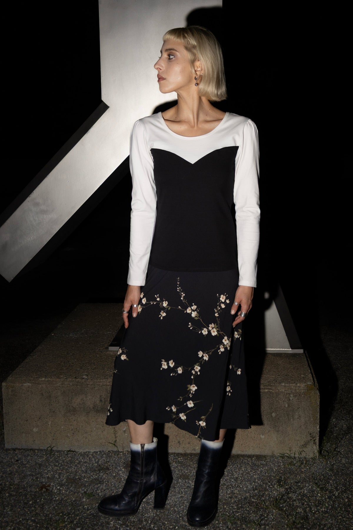 A woman wearing the Pearl Hart Top by Eve Lavoie, with a colour-blocked sweetheart neckline effect in black and white, and long sleeves with a puff at the shoulders. She is wearing it with a floral skirt and standing outdoors by a pillar at night. 