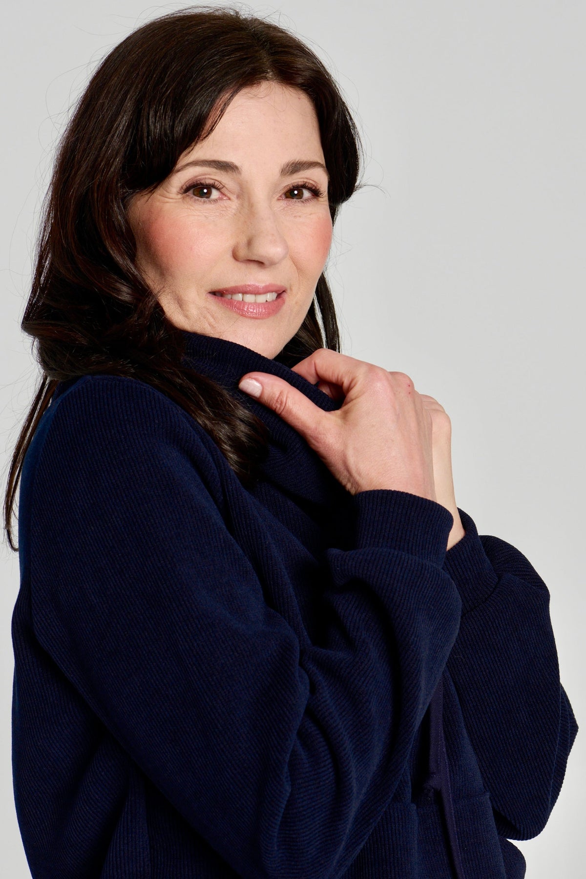 Close up side view of a woman wearing the Ramos Sweater by Kollonta in a ribbed navy corduroy-like fabric. The Sweater has a large cowl neck with a drawstring and a kangaroo pocket. She is standing against a white background.