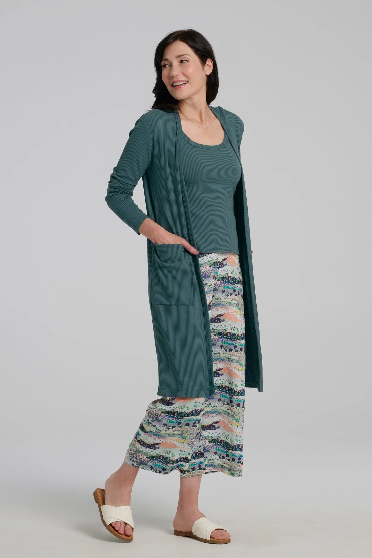 A woman wearing the Streliski Cardigan from Kollontai in Teal, a long open cardigan in bamboo rib with large patch pockets. She is wearing it with a matching tank and patterned pants on a plain background