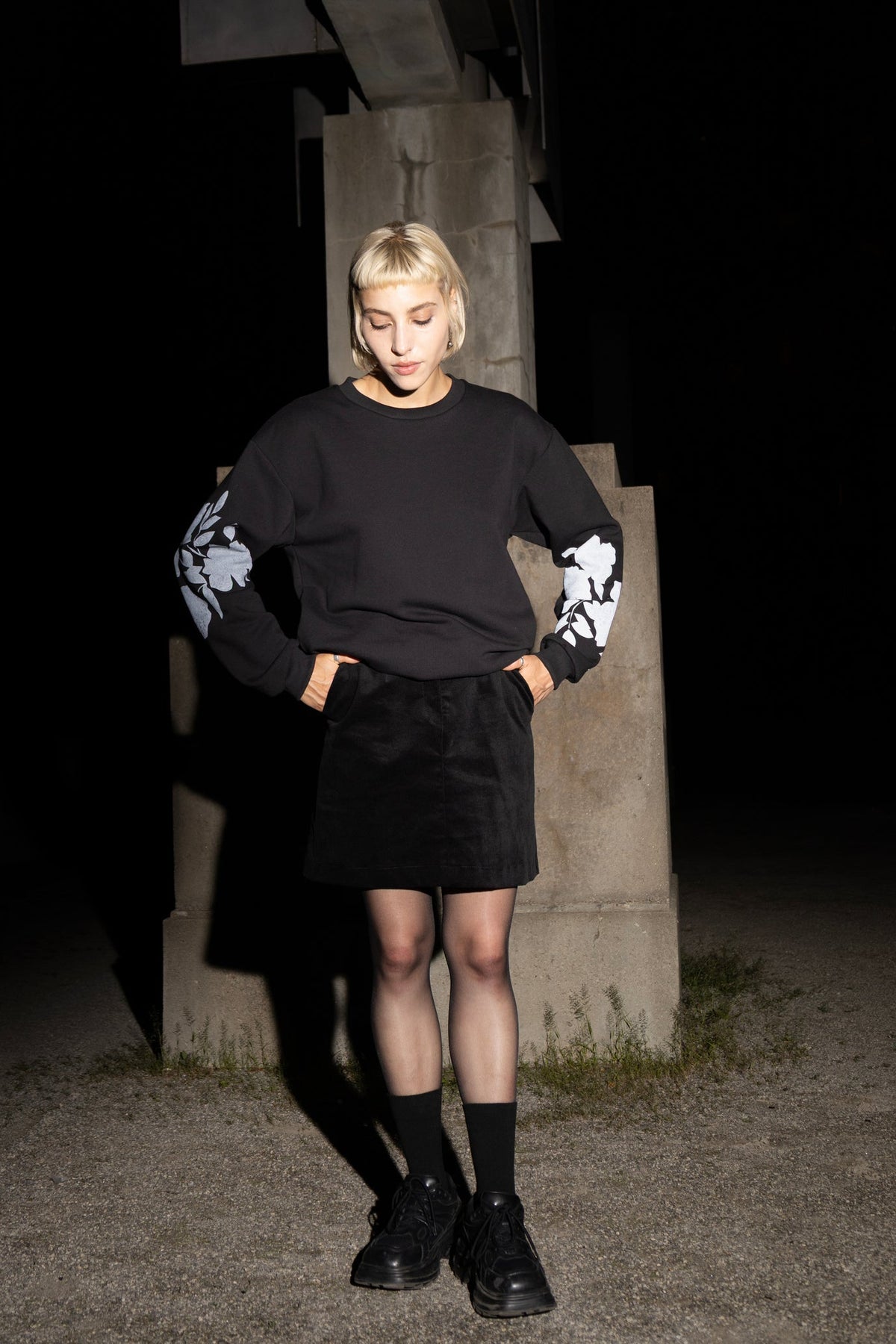 A woman wearing the Screen-Printed Crewneck Sweater by Eve Lavoie in Black with white floral patterns on the sleeves, paired with a black skirt, standing outdoors at night next to a concrete pillar.