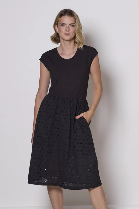 A woman wearing the Primrose Dress by Tangente in Black, a below the knee length dress featuring a bamboo rayon bodice and daisy eyelet skirt, with short sleeves and a round neck, standing against a plain background.