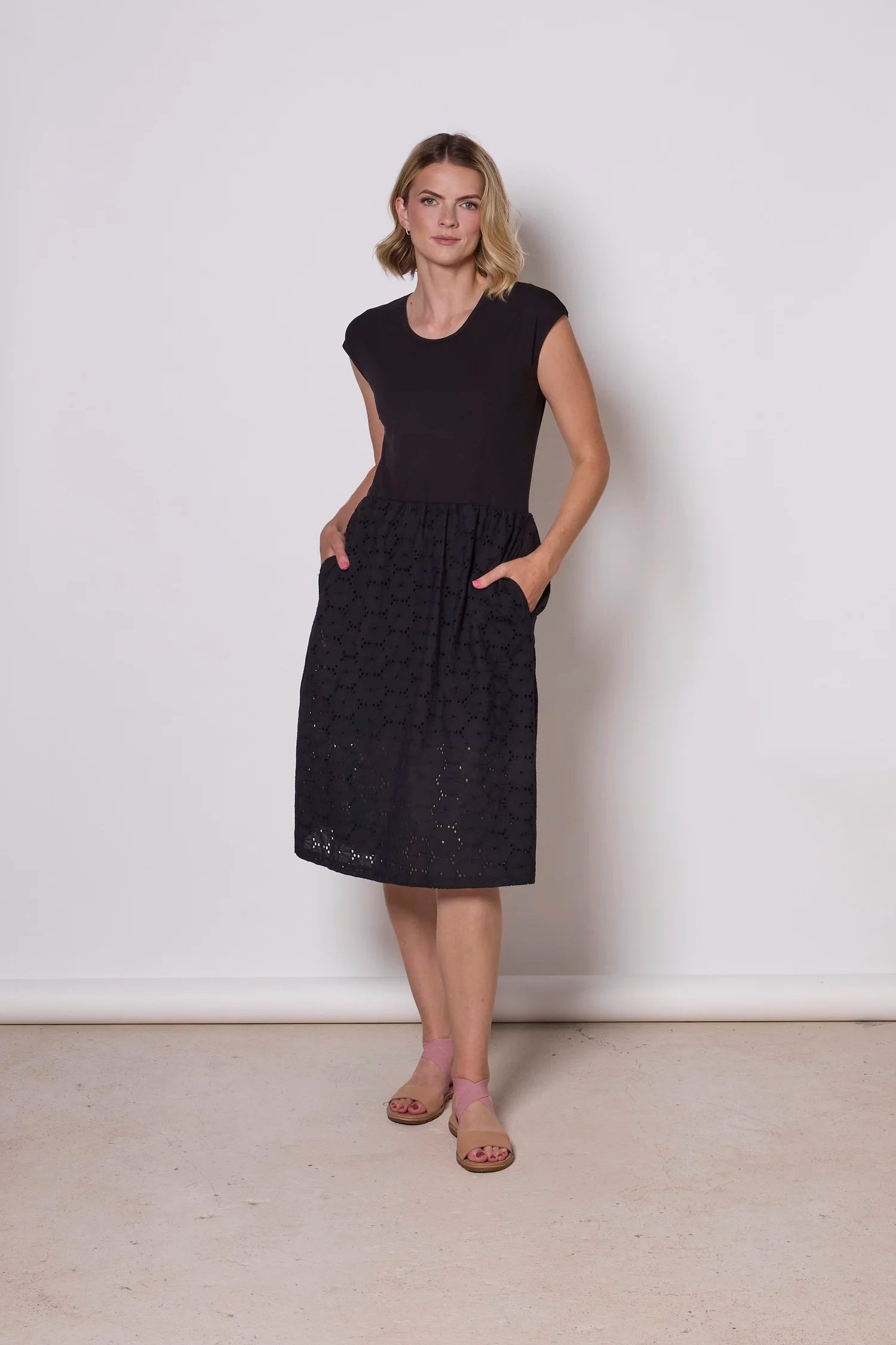 A woman wearing the Primrose Dress by Tangente in Black,  a below the knee length dress featuring a bamboo rayon bodice and daisy eyelet skirt, with short sleeves and a round neck, standing against a plain background. 