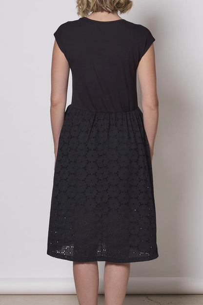 Back view of a woman wearing the Primrose Dress by Tangente in Black, a below the knee length dress featuring a bamboo rayon bodice and daisy eyelet skirt, with short sleeves and a round neck, standing against a plain background. 
