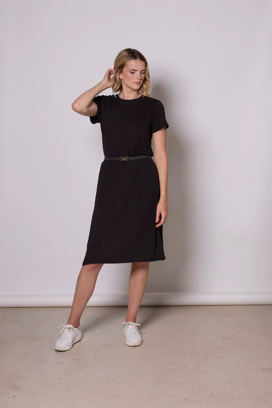 A woman wearing the Frances Midi  by Tangente in Black, a short sleeved loose fitting dress with side slits. She is wearing it with a black belt and standing against a plain background
