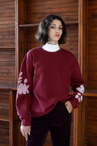 A woman wearing the Screen-Printed Crewneck Sweater by Eve Lavoie in Burgundy with white floral patterns on the sleeves, paired with purple pants, standing indoors by a wooden wall.