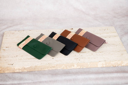 Five Leather Wallets from Armoir are arranged in a fan shape on a light beige stone slab over a soft light fabric background. 