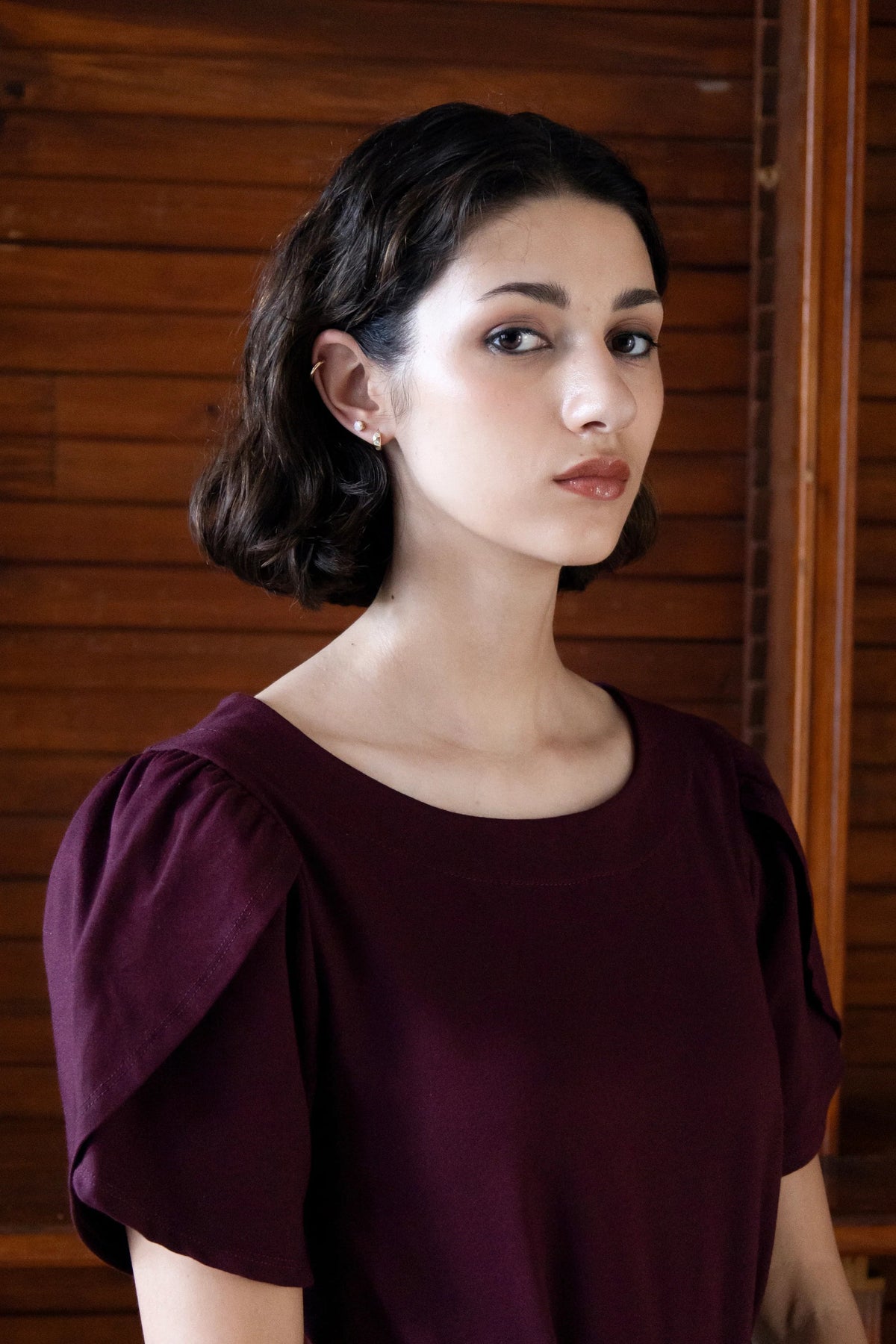 A woman wearing the Tanzania Tulip Top by Eve Lavoie in Burgundy, with a round neck and tulip sleeves. She is standing indoors by a wood panelled wall.