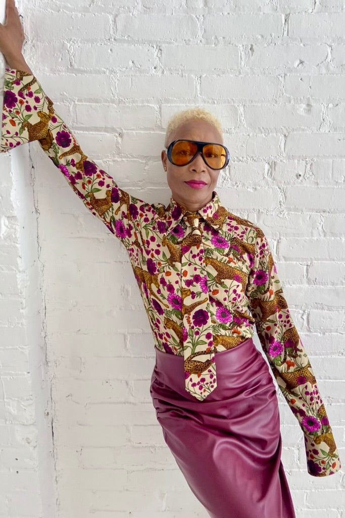 A woman wearing the Untamed Bloom Shirt by Dotty, a menswear-inspired blouse with a floral and animal print, classic button front details, and polished gold buttons. She is wearing it with a pink pleather skirt and leaning against a white brick wall. 
