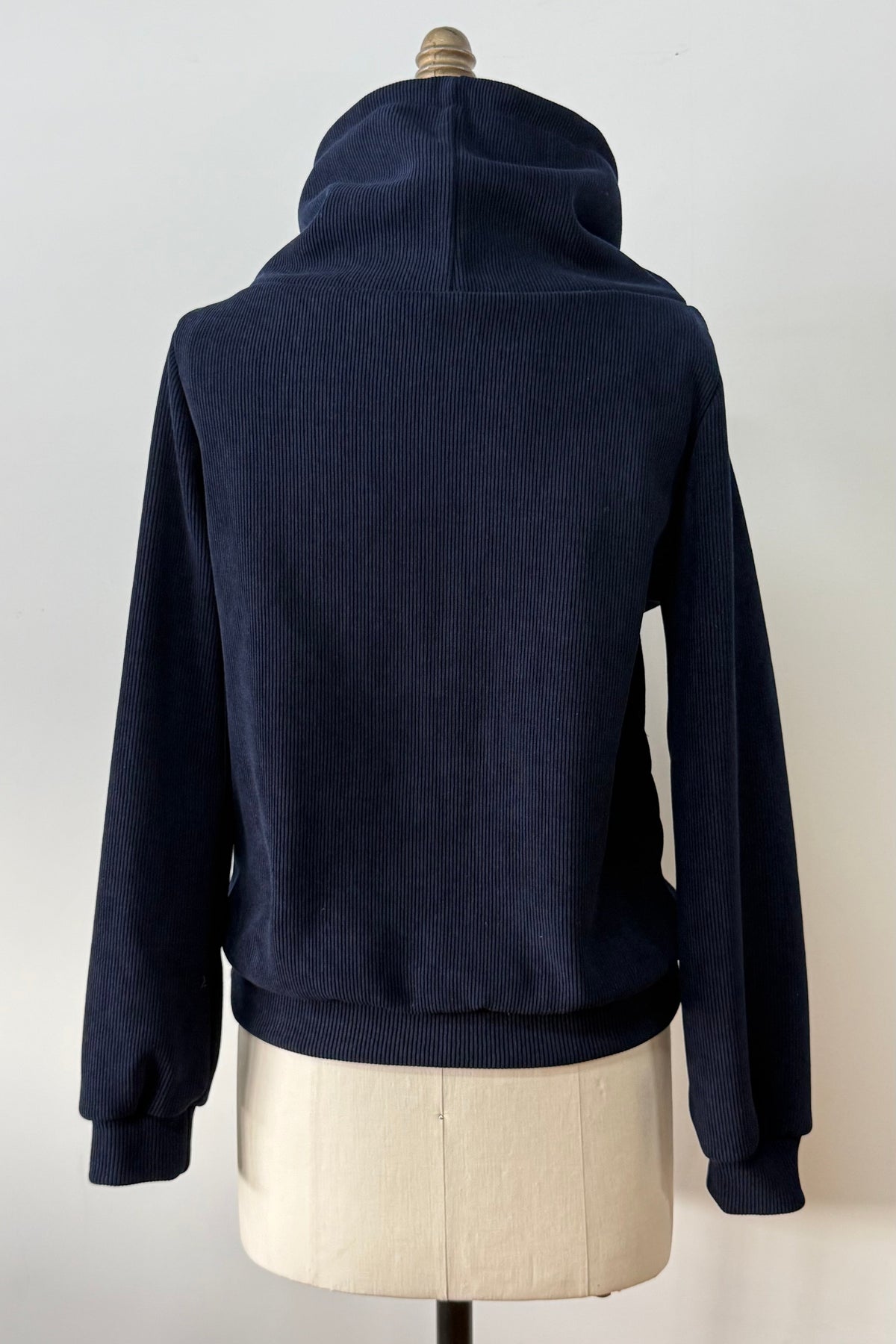A back view of the Ramos Sweater by Kollontai in a ribbed navy corduroy-like fabric, with a large cowl neck with a drawstring and a kangaroo pocket, is shown on a mannequin against a white background. 