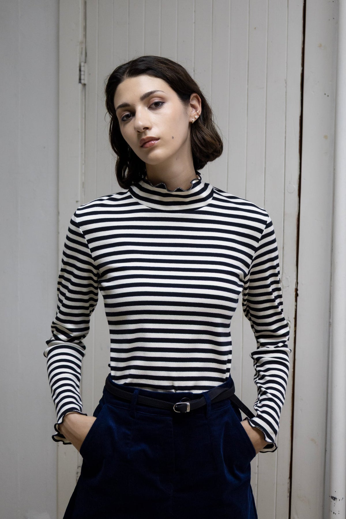 A model stands against a dark concrete pillar wearing the High-necked Moonstone Top from Eve Lavoie in Black and White Stripes with the Onyx mini skirt. She is standing in front of a white door. 