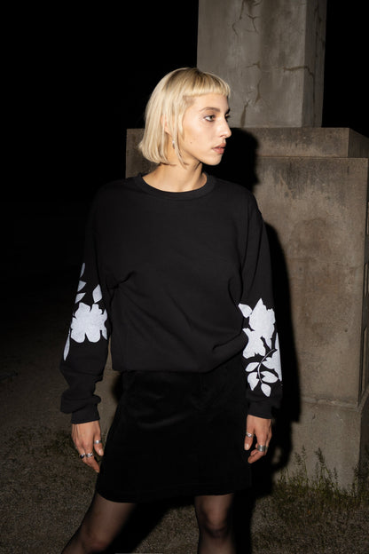 A woman wearing the Screen-Printed Crewneck Sweater by Eve Lavoie in Black with white floral patterns on the sleeves, paired with a black skirt, standing outdoors at night next to a concrete pillar.