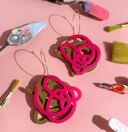 Pair of pink and green earrings on a pink background with art supplies.