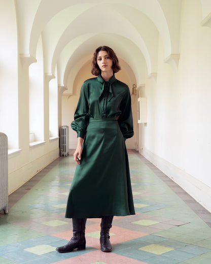 A woman wearing the Crystal Long Skirt by Eve Lavoie in Green Satin, an A-line maxi skirt. She's wearing it with a matching blouse and standing a long white hallway. 