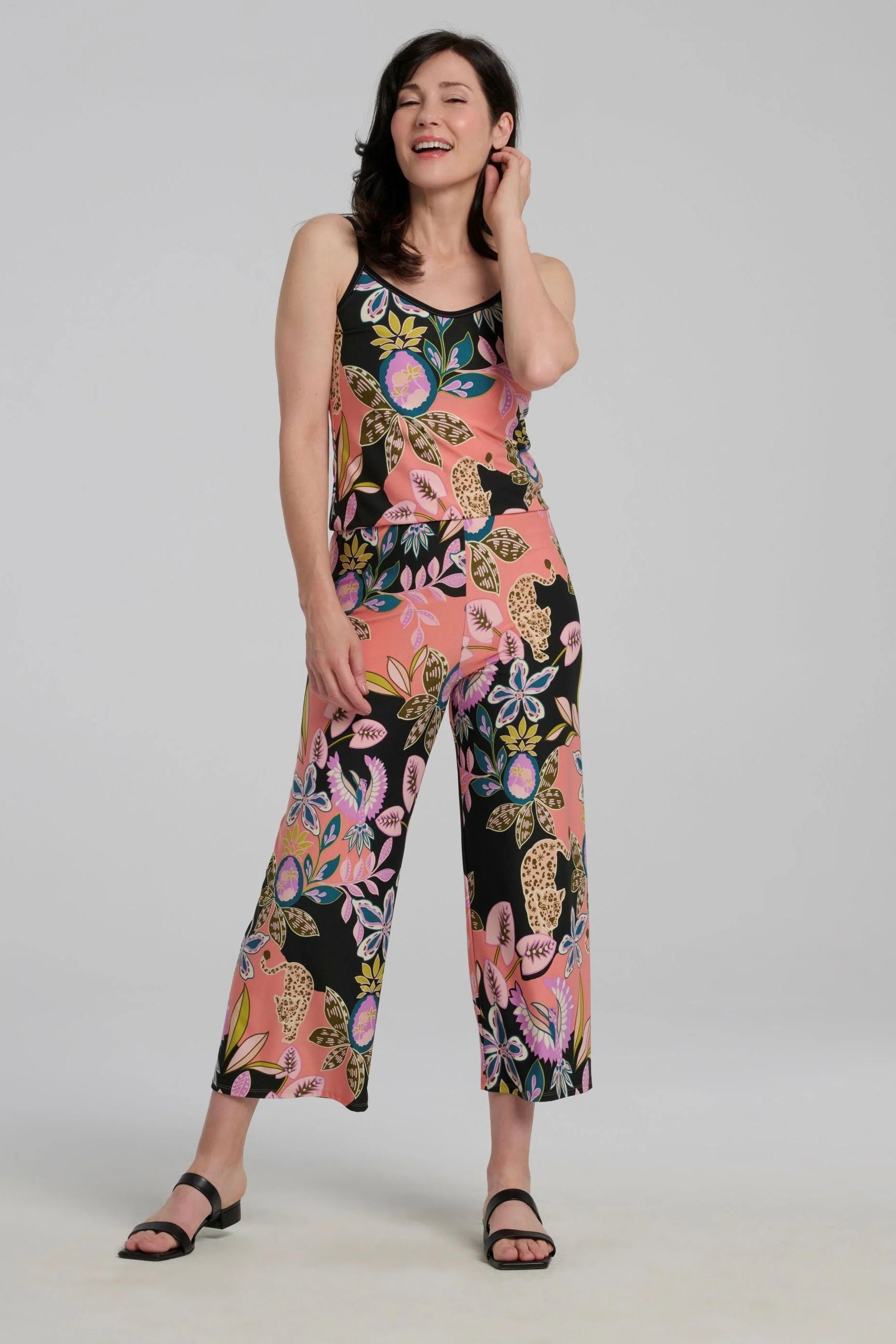 A woman wearing the Ortiga Jumpsuit by Kollontai, featuring a cheetah/floral print, spaghetti straps, an elastic waist, and 7/8 straight legs. She is standing in front of a grey background.
