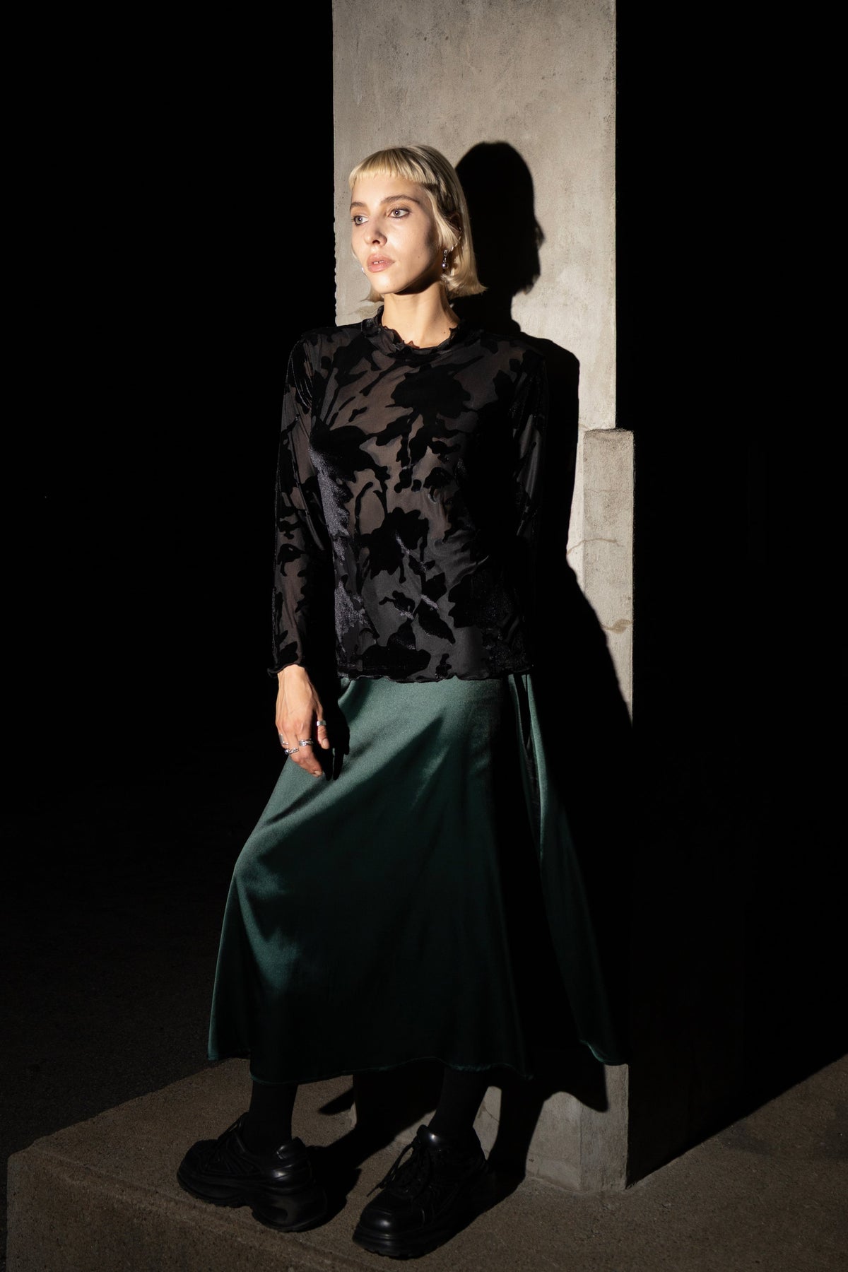 A model stands against a dark concrete pillar wearing the High-necked Moonstone Top from Eve Lavoie in Black Lace, with the Crystal skirt in Green satin. 