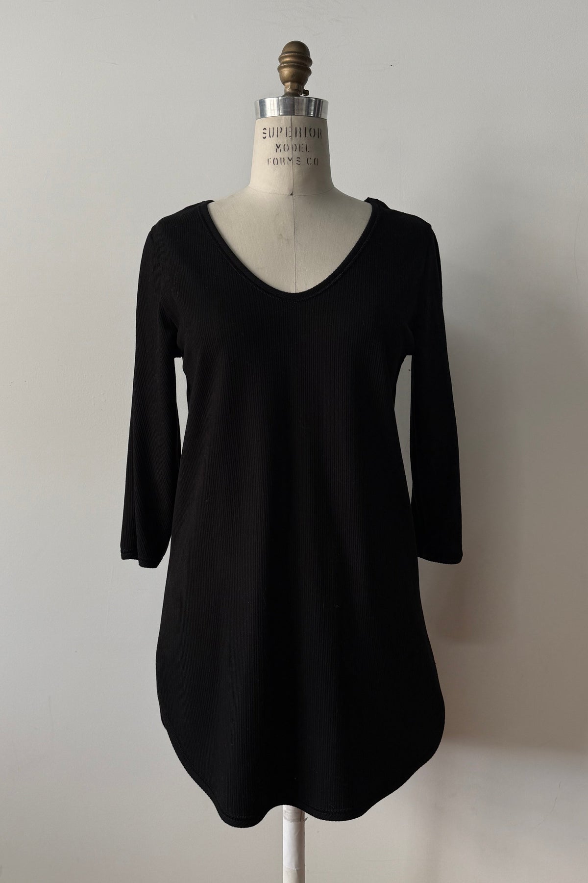 The Clara Tunic by Kollontai in Black, a stretch corduroy piece with a V-neck, a rounded hem, 3/4 sleeves, and inverted pleats at the shoulders, is shown on a mannequin against a grey background. 