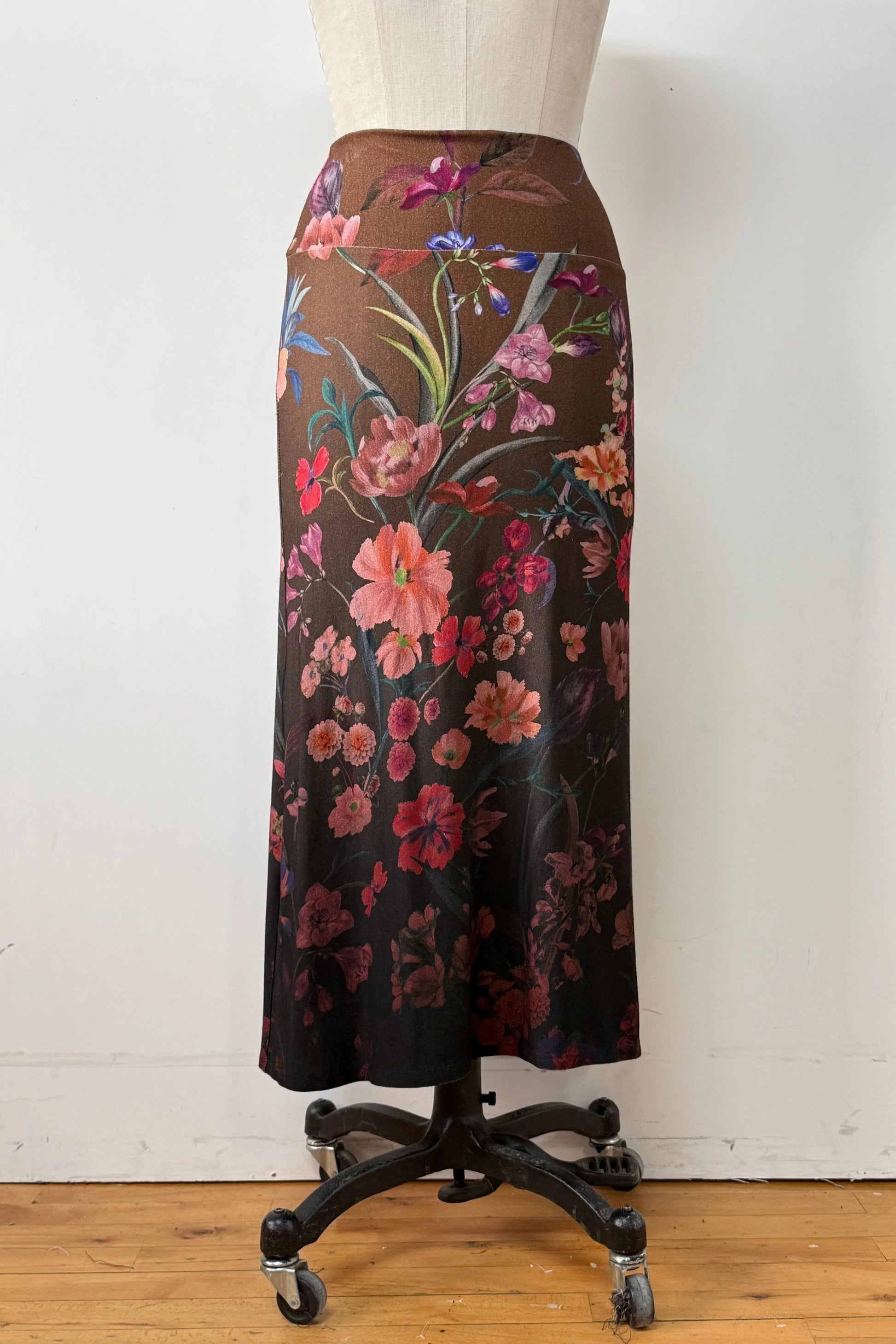 A back view of the Charlotte SKirt by Kollontai, a long slim cut brown skirt adorned with a floral pattern, is shown on a mannequin against a white background. 