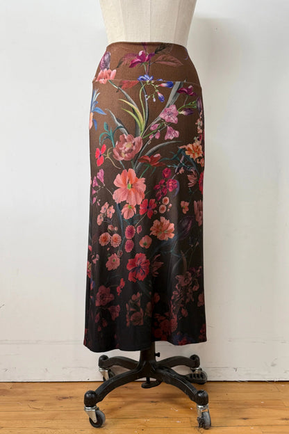 A back view of the Charlotte SKirt by Kollontai, a long slim cut brown skirt adorned with a floral pattern, is shown on a mannequin against a white background. 