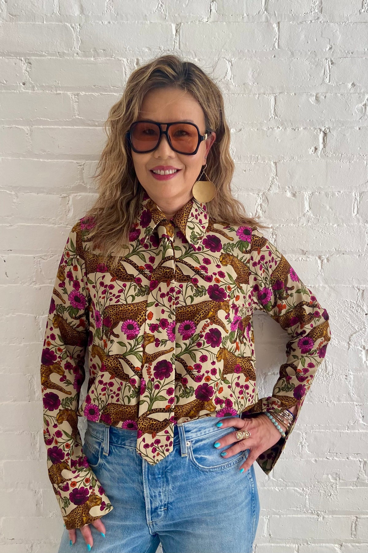 A woman wearing the Untamed Bloom Shirt by Dotty, a menswear-inspired blouse with a floral and animal print, classic button front details,, and polished gold buttons. She is wearing it with jeans and leaning against a white brick wall. 