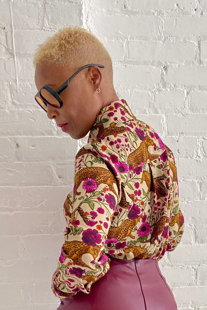 Side view of a woman wearing the Untamed Bloom Shirt by Dotty, a menswear-inspired blouse with a floral and animal print, classic button front details, and polished gold buttons. She is wearing it with a pink pleather skirt and leaning against a white brick wall. 