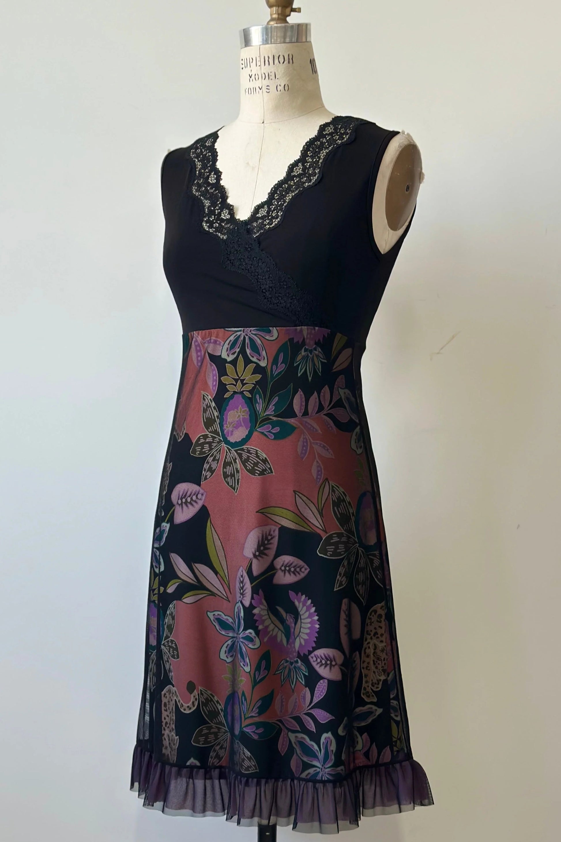 The Liseron Dress by Kollontai is shown on a mannequin against a grey background. It's a sleeveless dress with a faux-wrap neckline, empire waist, knee-length a-line skirt. The bodice is solid black, while the skirt features a cheetah/floral print under a mesh overlay. 