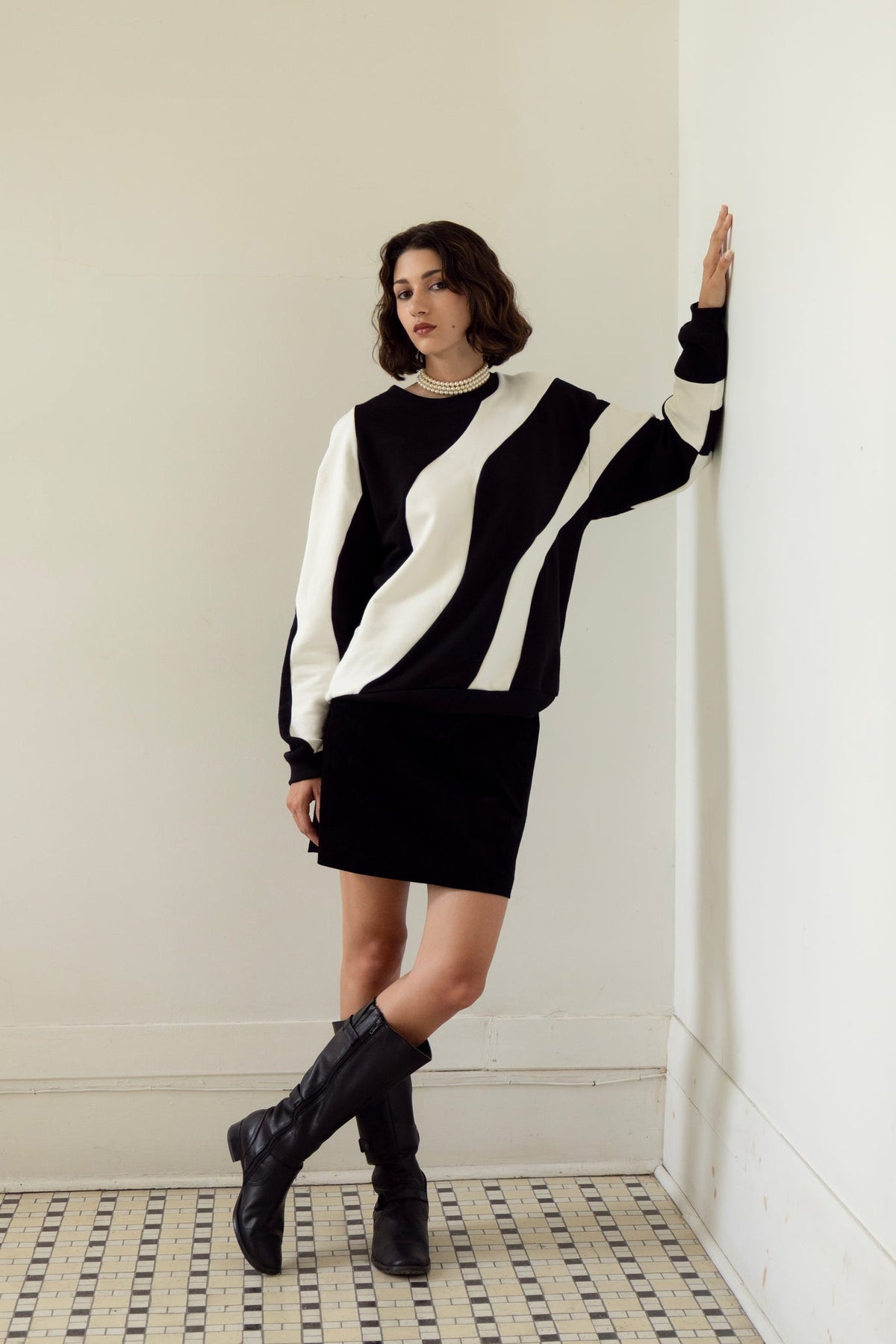 A woman wearing the Amber Sweatshirt by Eve Lavoie in Black/Ivory, an oversized unisex sweatshirt with wave-shaped panels. She is wearing it with a black miniskirt and standing in a white room with a patterned floor.