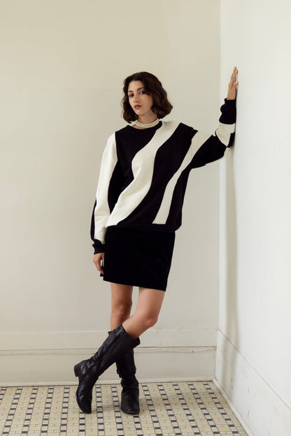 A woman wearing the Amber Sweatshirt by Eve Lavoie in Black/Ivory, an oversized unisex sweatshirt with wave-shaped panels. She is wearing it with a black miniskirt and standing in a white room with a patterned floor.