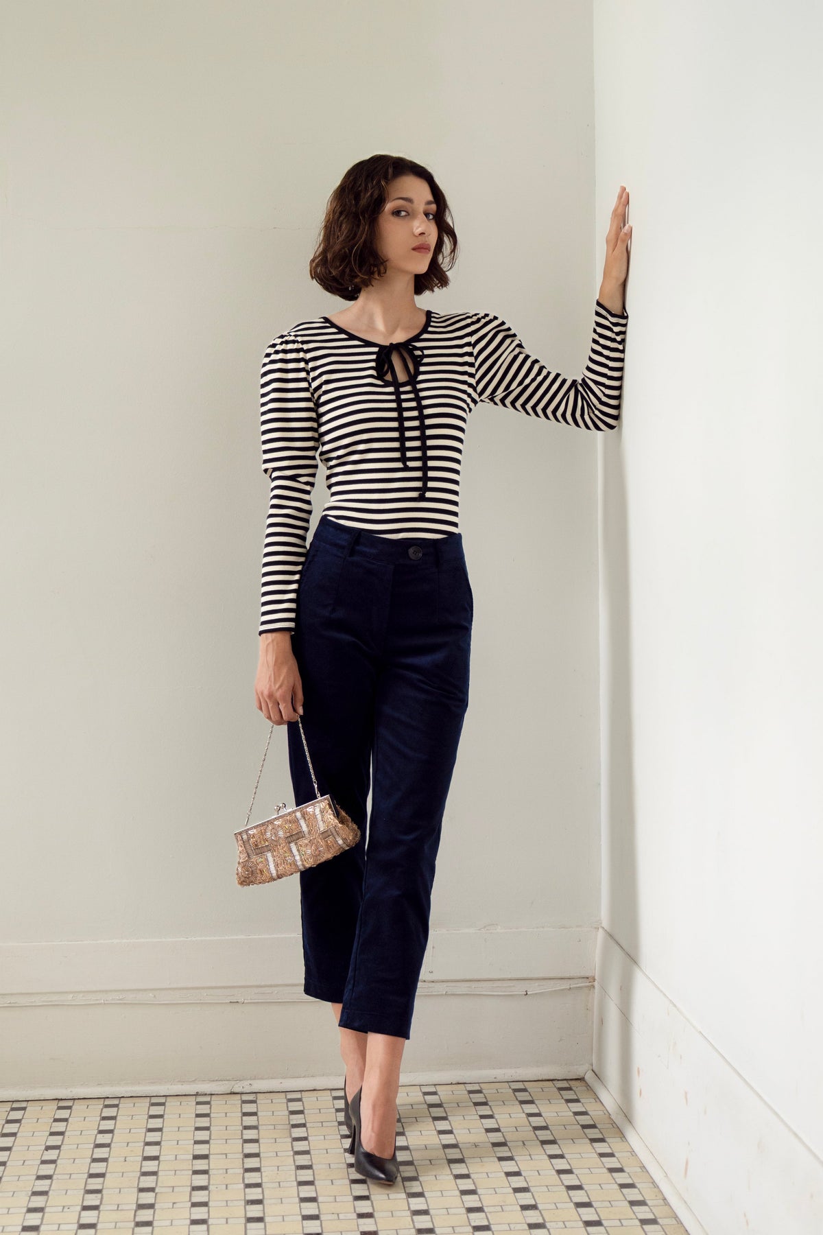 A woman wearing the Cornaline Top by Eve Lavoie, a reversible black and white striped long-sleeve top with a cut-out and tie neckline on one side and a round neckline on the other, plus puff sleeves. She is wearing it with navy cropped pants and standing in a white room. 