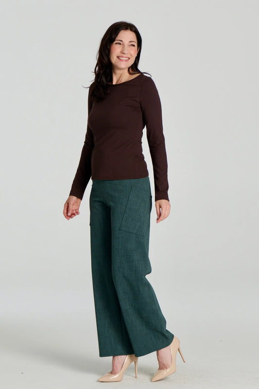 A woman wearing the Loretto Top by Kollontai in Chocolate, a slim fitting long sleeved top with a round neck. She is wearing it with the Susan pants in Green and standing in front of a grey background. 