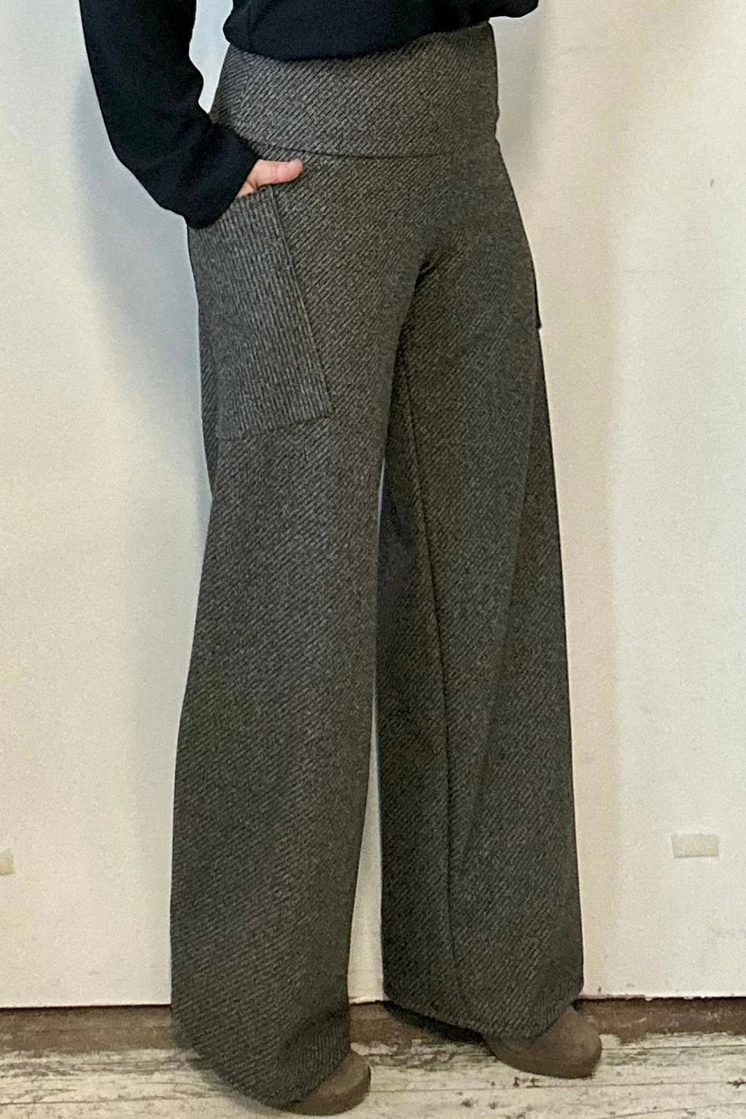 Waist down view of a woman wearing the Susan Pants by Kollontai in Charcoal, wide pants with a wide pull on waistband and angled patch pockets. She is wearing them with a black top and standing in front of a white wall. 