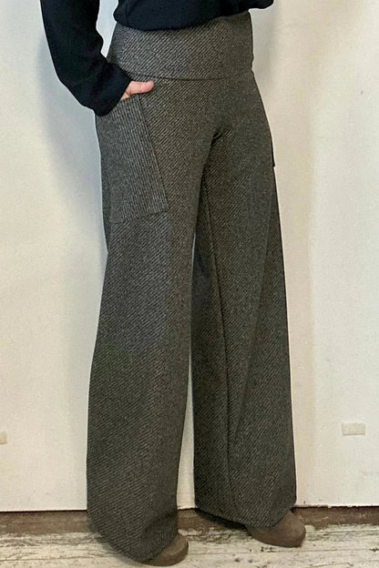 Waist down view of a woman wearing the Susan Pants by Kollontai in Charcoal, wide pants with a wide pull on waistband and angled patch pockets. She is wearing them with a black top and standing in front of a white wall. 
