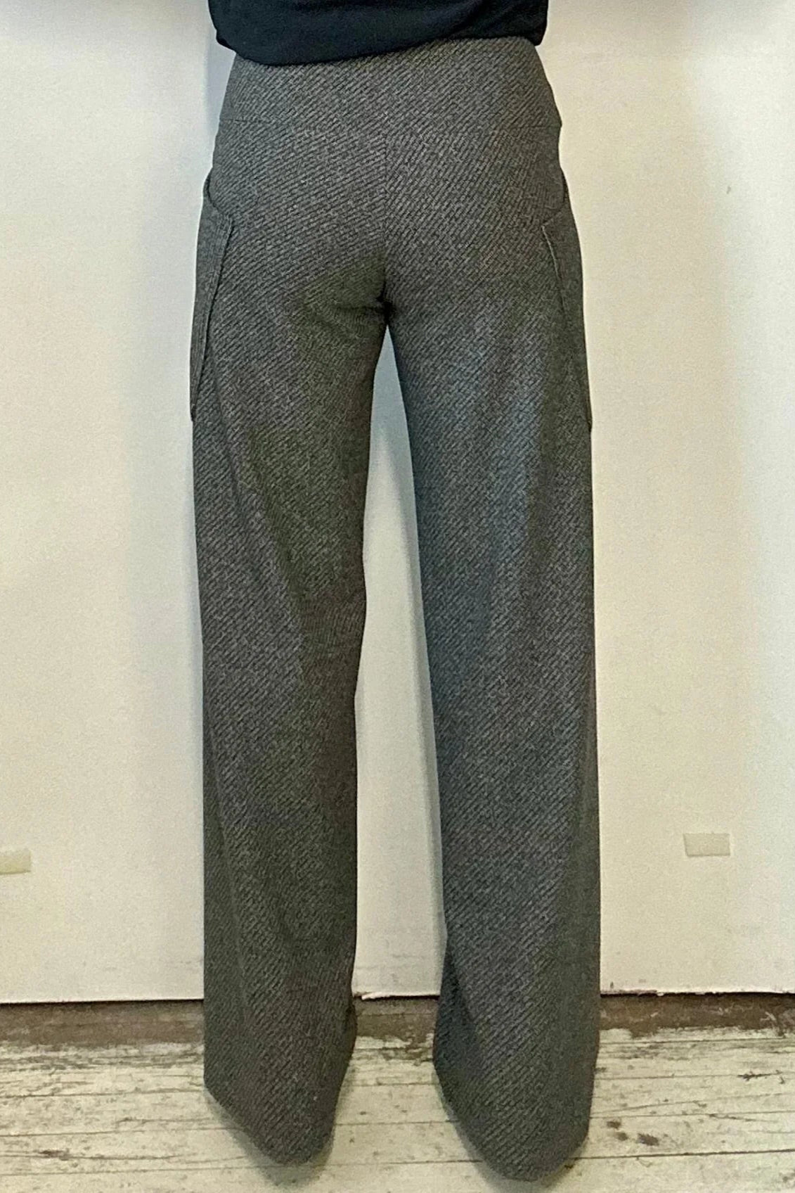 Back waist down view of a woman wearing the Susan Pants by Kollontai in Charcoal, wide pants with a wide pull on waistband and angled patch pockets. She is wearing them with a black top and standing in front of a white wall. 