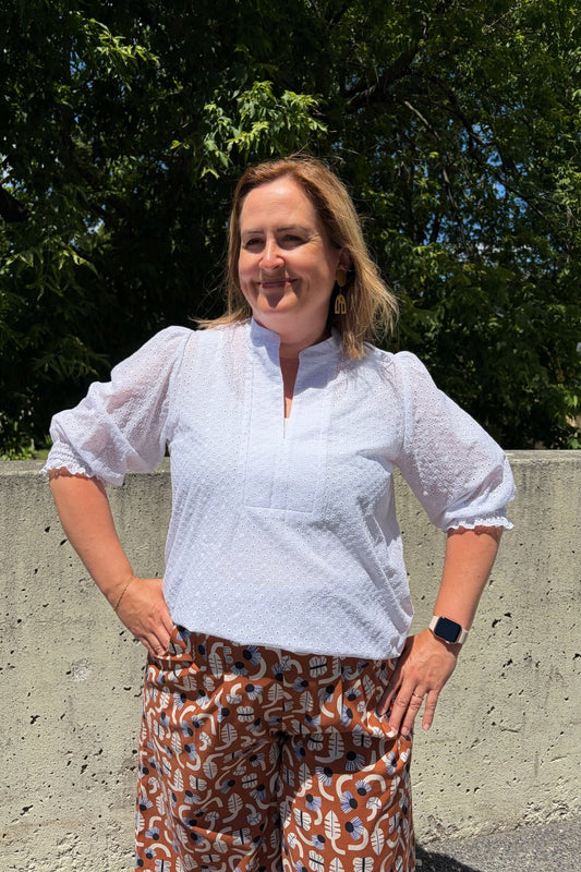 A woman wearing the Eyelet Summer Blouse by Dotty in White, a pullover top with a banded v-neck and 3/4 smock sleeves. She is wearing it with patterned pants and standing outside by a cement wall. 